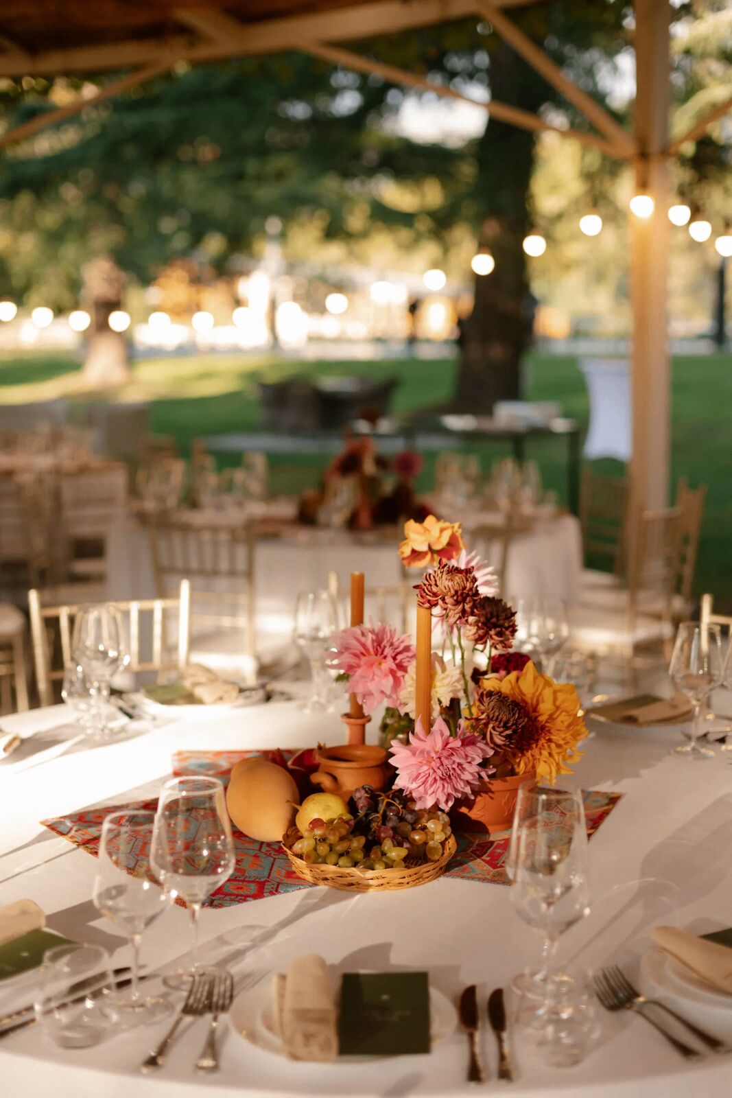 Open-air garden dinner with dahlia and fruit centrepiece, taper candles and string lights at golden hour — a Well Done Events tablescape