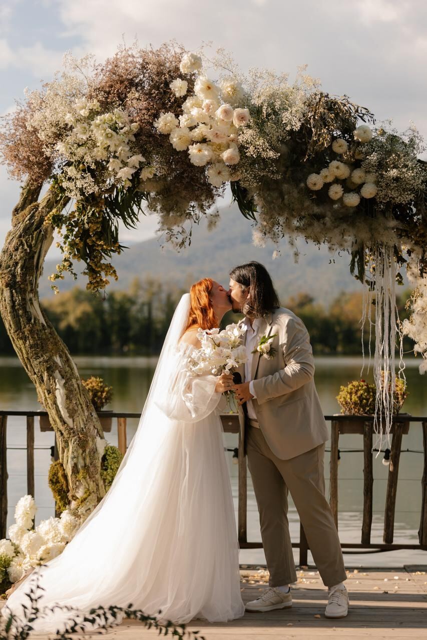 Bride and groom kissing under a floral arch by the lake in Kakheti